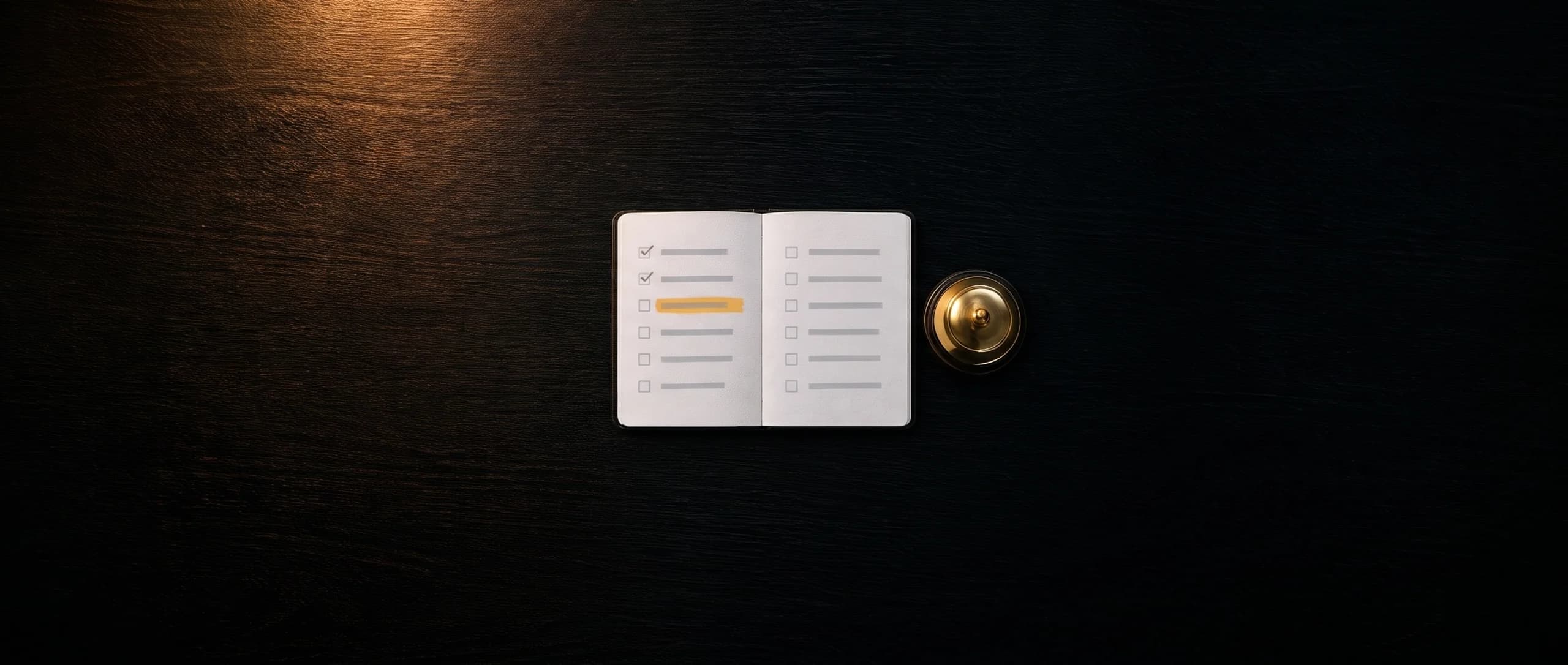 Ein aufgeschlagenes Notizbuch mit einer Liste, zwei Punkte abgehakt, einer in Amber markiert, daneben eine Tischglocke aus Messing, auf dunklem Holz